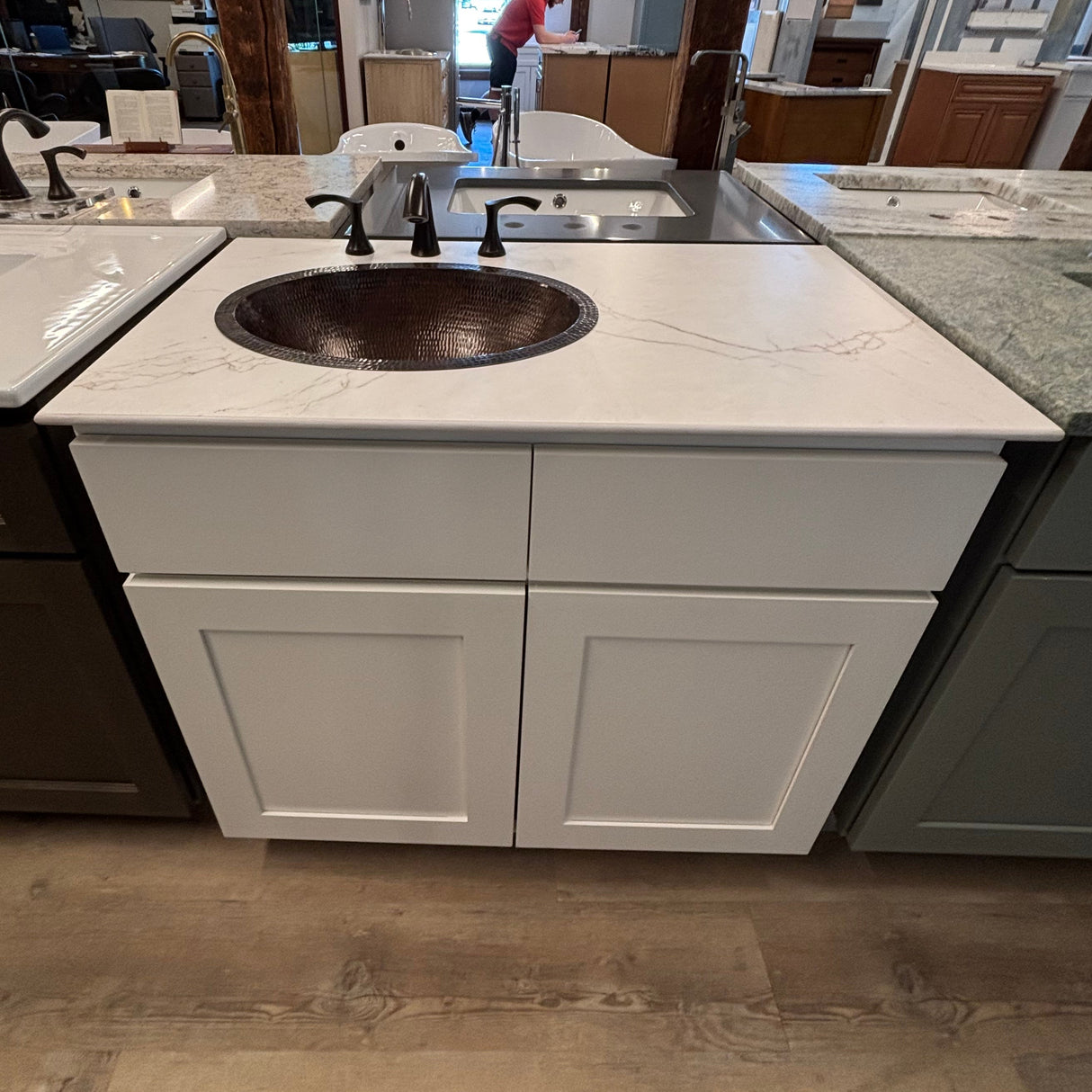 Kitchen showroom with white cabinets, granite countertops, and a person in the background.